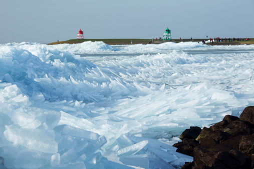 Ice formation, pushed up against a dyke by strong winds. Red and green harbor lights in the back. Unrecognizable people on the dyke in the back.