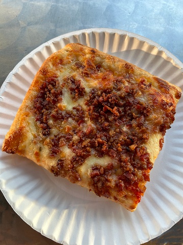 A corner square slice of pizza - sometimes called Sicilian style - and topped with tomato sauce, cheese, and bacon bits served on a white paper plate on a silver table top in a Hoboken, New Jersey pizzeria.