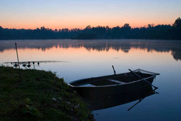 Back lit fishing rods and a boat at rest. Colors of dawn in the clear romantic sky over the woods in the background, mirrored on the water. Long shutter speed for smooth water surface. Photographed at a calm lake in Vendeuil, Picardy, France, Europe.