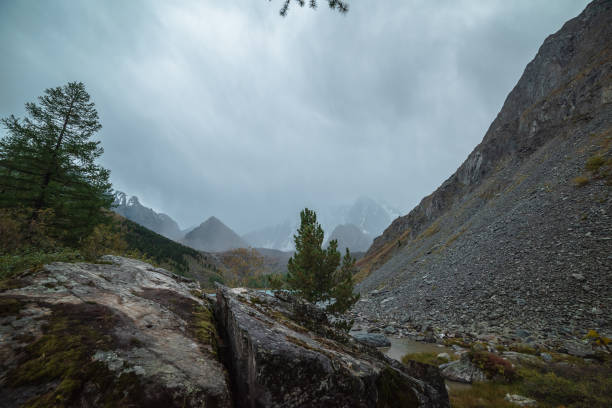 istock Mossy cracked rock among coniferous trees against pyramid shaped mountain silhouette during rain in overcast. Gloomy view to crack stone and snowy mountains under gray cloudy sky. Fading autumn colors 1606317638