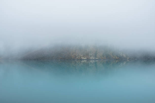 istock Simple meditative landscape with turquoise lake in thick fog. Tranquil view through clearance in dense fog to steep slope in fading autumn colors and mirror mountain lake. Reflective azure alpine lake 1606317640