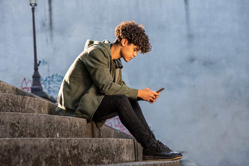 Full body profile portrait young man sitting on steps with mobile phone