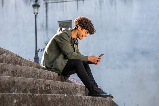 Full body profile portrait happy young man sitting on steps with mobile phone