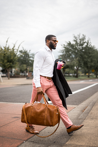 Young, handsome man of African American ethnicity wearing business casual clothing and sunglasses walks across the street carrying a brown, leather duffel bag, jacket and an iced drink.