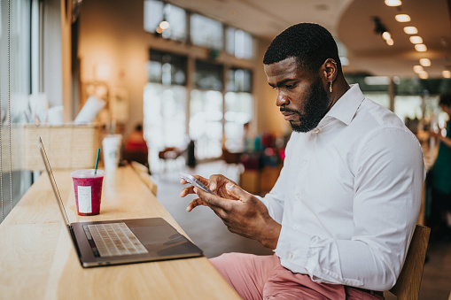 Young man of African American ethnicity wearing business casual clothing glances at his phone while working on his laptop with an iced drink in a busy cafe.