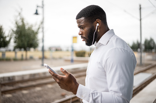 Well-dressed black businessman wearing headphone and using his phone while waiting on train station platform during commute to work.