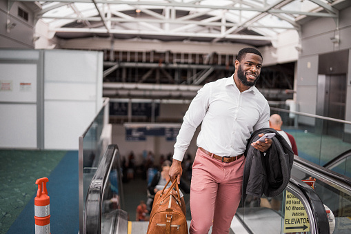 Smiling African American businessman on a work trip carries a duffle bag while riding up an escalator in an airport.