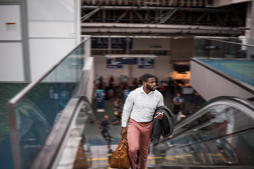 African American businessman on a work trip carries a duffle bag while riding up an escalator in an airport.