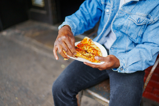 Cropped view of an unrecognizable black man holding a slice of pizza while dining outside at casual restaurant.