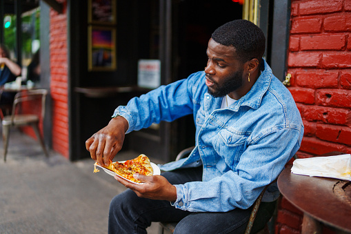 Hip young black man enjoying a slice of pizza outside a casual restaurant in a trendy downtown neighborhood.