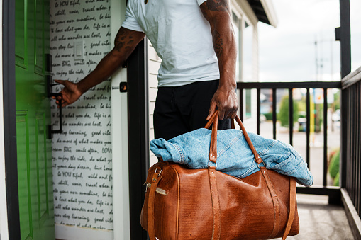 Cropped view of an unrecognizable black man who is leaving on a trip carrying a brown leather duffel bag as he walks out the front door of his home.