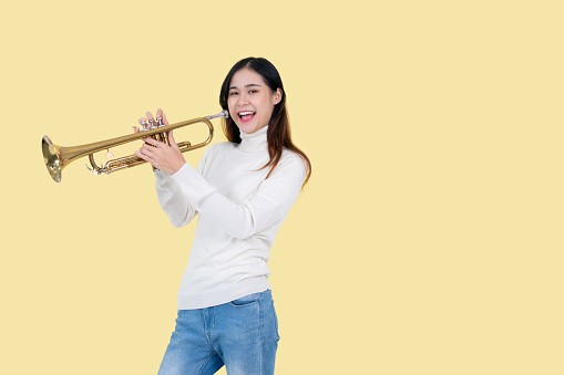 A playful and talented Asian female musician with a trumpet stands against an isolated yellow background. Leisure activity, music school student, hobby