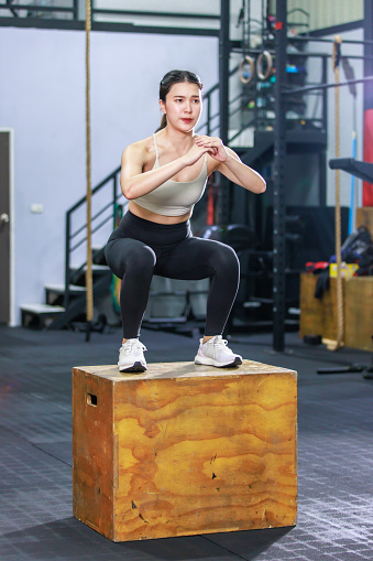 Millennial Asian fit strong muscular female fitness model athlete in sport bra legging and sneakers working out training exercising squatting on wooden box alone in gym full of exercise equipment.