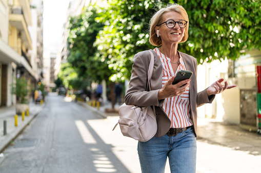 Smiling mature businesswoman talking on smart phone with wireless headphones in ears while walking in the city