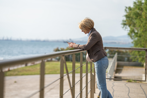 Mature businesswoman checking emails on mobile phone while leaning on promenade fence in coastal town