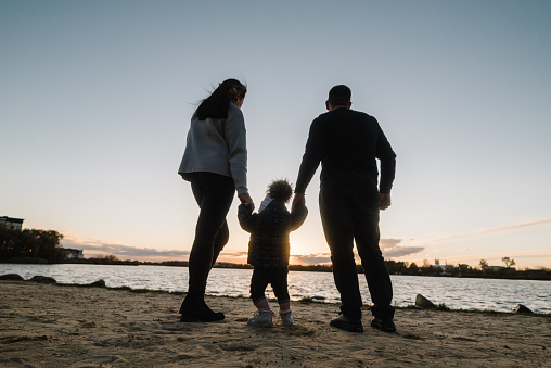 Silhouette at sunset. Mother, father and daughter near lake in nature. Spending time together on beach on vacation. Mom, dad hugs child, walk in park. Concept of autumn holiday. Family photo outdoors