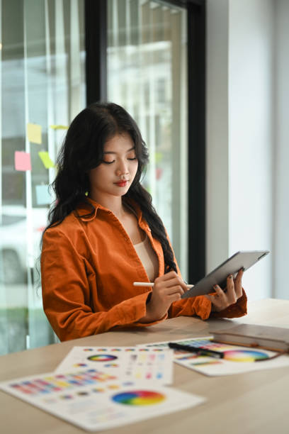 imagen de una elegante mujer asiática con chaqueta naranja usando una tableta digital, trabajando en una oficina creativa - fashion palette fotografías e imágenes de stock
