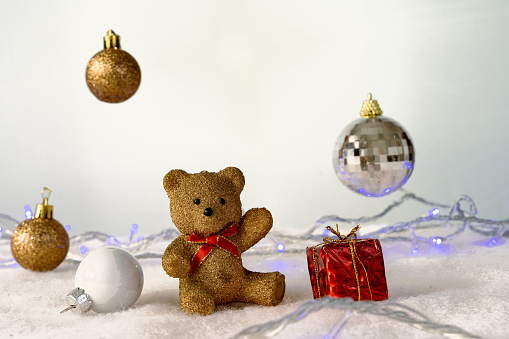 On a white background surrounded by Christmas decorations and lights, a funny little bear greets the camera with one hand, the bear is a Christmas ornament.