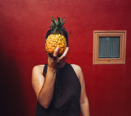 A woman plays with a pineapple in front of a red wall background