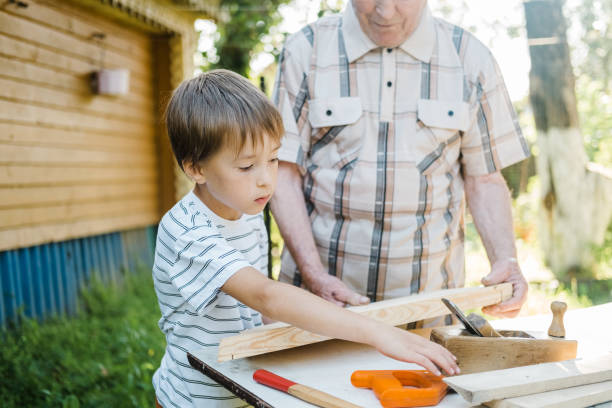 istock Elderly gray-haired mature man and a small boy are standing at a table with carpentry tools. Grandfather teaches his grandson to carpenter outdoors in the backyard of the house on a sunny day. 1607109973