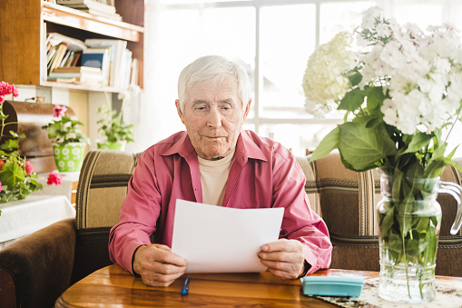An elderly Caucasian man with gray hair is sitting at home in the living room and studying financial documents, paying bills