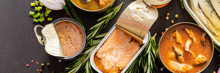 Different open tin cans with canned fish among spices and herbs banner, canned salmon and mackerel, sprat and sardine, tuna and herring and fish pate, top view