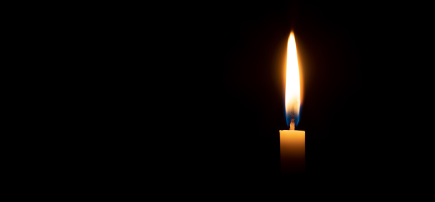 Single burning candle flame or light is glowing on a small white candle on black or dark background on table in church for Christmas, funeral or memorial service with copy space.