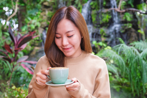 Portrait image of a young woman holding and drinking hot coffee with nature in background