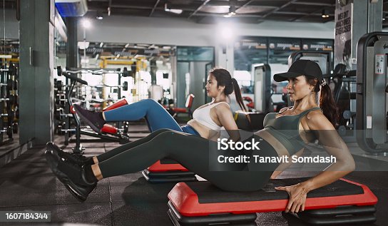 istock Two young woman doing knee tuck crunches in the gym. Powerful female bodybuilder pumping abs muscles. 1607184344