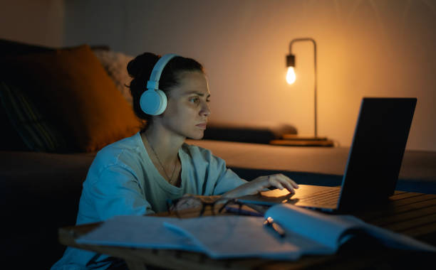 Young caucasian female student in glasses and headphones working on a project at home at night using a laptop. Work and education online and deadline concept stock photo