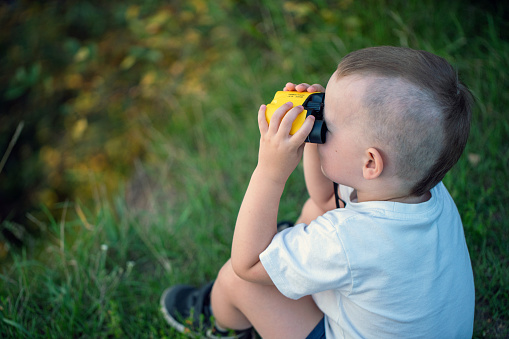 Cute boy looking through binoculars while sitting on the grass, back top view