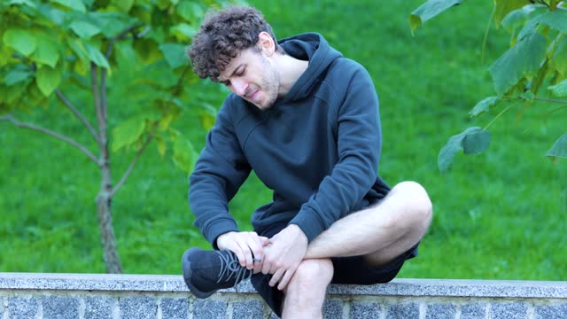 Caucasian man sitting and massaging his leg in green park. Sportsman feeling strong pain in his ankle and foot. Handsome man with brown curly hair starching his leg.