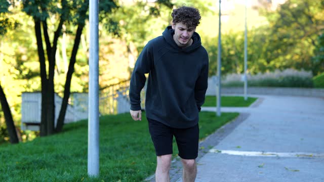 Young Caucasian man feeling pain after working out. Sportsman running in park, feeling leg hurt after outdoor activities. Face of guy showing strong pain.