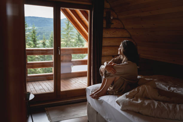 junge kaukasische frau sitzt auf einem bett in einer hölzernen chalethütte in den bergen. komfortables ausruhen und schlafen in einem blockhaus in der natur - rustic bedroom cabin indoors stock-fotos und bilder