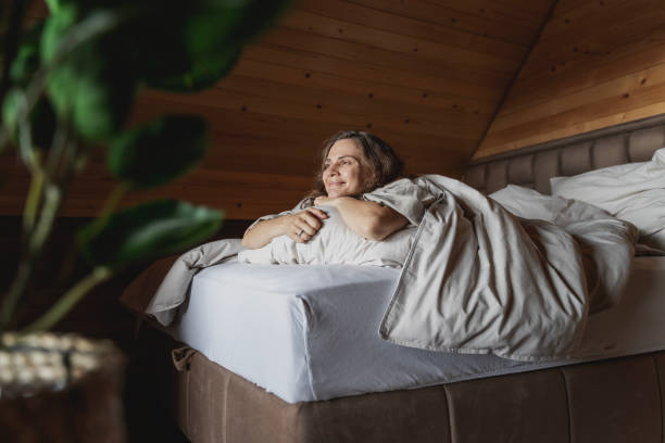 young happy caucasian woman lying on bed with beige pastel linens hugging pillow and enjoying comfortable rest in log house - rustic bedroom cabin indoors imagens e fotografias de stock