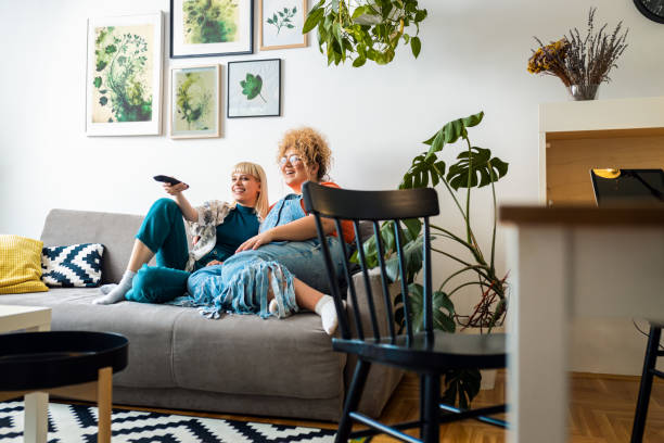 Smiling lesbian couple sit on the sofa, embracing each other and watching tv.