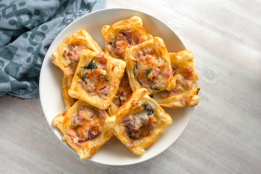 Baked puff pastry squares with ham, onion, herbs and cheese in a bowl, finger food snack for a warm or cold party buffet, high angle view from above, copy space, selected focus, narrow depth of field