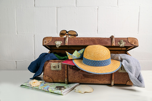 Vintage suitcase packed with travel accessories like straw hat, towels, passport, map and sunglasses for beach holidays in summer, rough white wall, copy space, selected focus, narrow depth of field