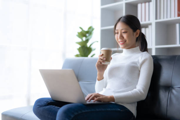 istock Happy young Asian woman relaxing using laptop computer and drinking coffee sitting on sofa at home. 1607362014