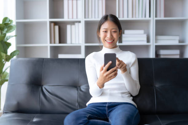 istock Beautiful young smiling Asian woman using smartphone on sofa at home. 1607362066