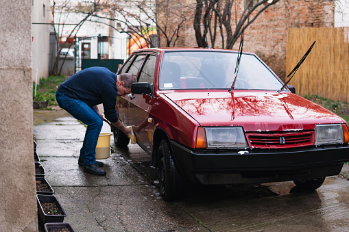 A senior man holds a sponge and washes a car in his backyard.
