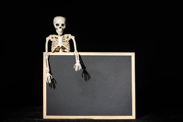 istock Funny human skeleton holding an empty black blank on a black background, copy space, mockup. Halloween Greeting Card 1607382404
