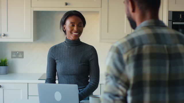 Couple At Home In Kitchen With Woman Working On Laptop Computer Talking With Man