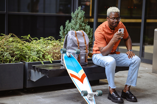 A young man sitting on a bench in the city and using a smart phone. His longboard is next to him
