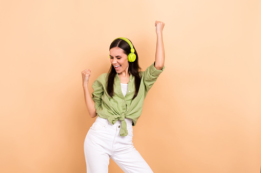 Photo of satisfied girl with straight hairstyle dressed green blouse in headphones fists up win lottery isolated on beige color background.