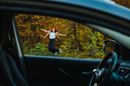 view through car window to road. woman in red beret jumps against background of autumn trees