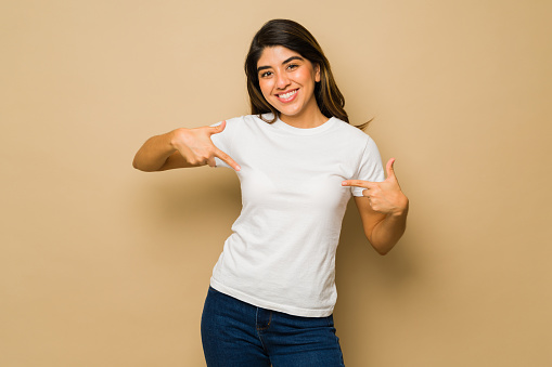 Attractive young woman pointing to her white mockup t-shirt smiling against a studio background