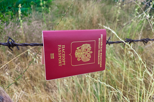Red passport of Russian Federation hangs on a rusty old barbed wire against the background of dry yellow grass. Concept of closing border, restricted area, isolation from the world. Some perspective