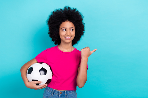 Photo of satisfied pleasant girl wear pink t-shirt holding soccer ball directing look empty space promo isolated on blue color background.