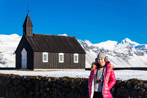 A young woman posing in Budakirkja or better known as The Black Church view with blue sky during winter snow, Iceland